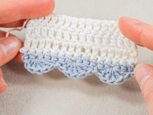 Gros plan de mains tenant un ouvrage au crochet avec une bordure coquille au crochet en fil bleu clair et des rangées de points blancs au-dessus, sur un fond de tissu clair.
