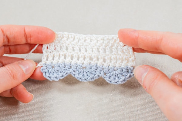 Deux mains tiennent un petit échantillon crocheté comportant une rangée blanche de points de double crochet sur le dessus et une bordure bleue coquille au crochet sur le dessous, présenté sur une surface claire.