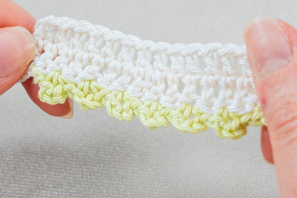Gros plan de deux mains tenant un morceau de crochet comportant des points blancs sur le dessus et une bordure vert clair coquille au crochet formant un bord décoratif en bas, sur un fond neutre.