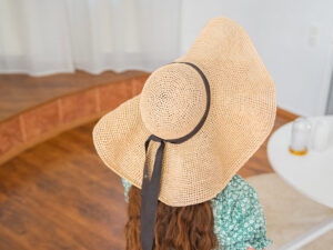 Une personne portant un grand chapeau au crochet avec un ruban noir est assise à l'intérieur sur un plancher en bois, de face. La personne a de longs cheveux bruns et est vêtue d'un haut vert à motifs.