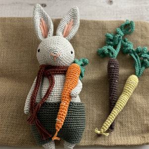 Un lapin en crochet portant un short vert et une écharpe marron tient une carotte orange en crochet. Deux autres carottes au crochet, l'une violette et l'autre jaune, sont posées à côté de lui sur une surface en toile de jute.
