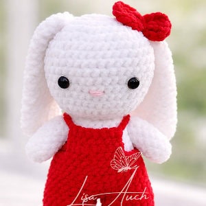 Un lapin blanc crocheté avec de longues oreilles, portant une robe rouge et un nœud rouge sur la tête. Le lapin a des yeux noirs en forme de boutons et une petite bouche rose. L'arrière-plan est légèrement flou.