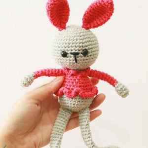 Une main tenant une poupée lapin crochetée avec de grandes oreilles roses, un corps gris, des yeux noirs et une tenue rose. Le lapin a de longues pattes molles et un petit visage simple.