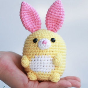 Un petit lapin en crochet jaune, fait à la main, avec des oreilles roses, des yeux noirs, un nez blanc et un ventre blanc, est tenu dans la paume de la main d'une personne.