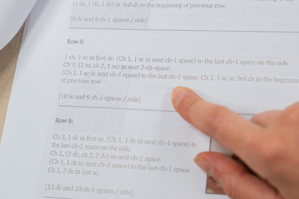 Une main désigne une ligne précise des instructions d'un modèle de crochet imprimées sur une feuille de papier, sur laquelle on distingue le texte, les rangs numérotés et un lexique anglais-français du crochet, pour faciliter la consultation.