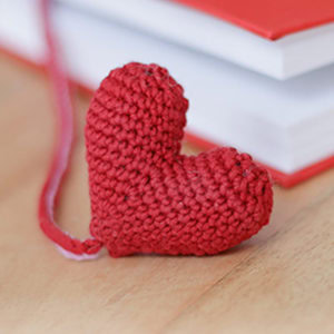 Un petit porte-clés rouge tricoté en forme de cœur repose sur une surface en bois à côté d'un livre fermé à la couverture rouge.