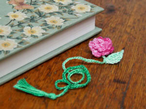 Une marque pages au crochet représentant une fleur rose, une feuille verte et un pompon est posée sur une table en bois à côté d'un livre à couverture rigide avec un motif floral.