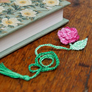 Un livre à motifs floraux est posé sur une surface en bois, fermé, avec un marque-page en crochet rose et feuilles vertes partiellement glissé entre ses pages.