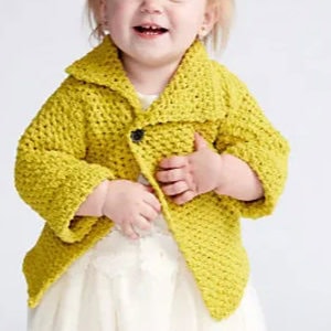 Un bambin souriant portant une veste en crochet jaune texturée sur une robe blanche, debout sur un fond clair uni.