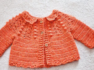 Un pull-over orange au crochet pour bébé avec des manches longues, un motif texturé, un petit col et deux boutons en bois sur le devant, posé à plat sur une surface texturée légère-idéal comme modèle gratuit de brassière au crochet.