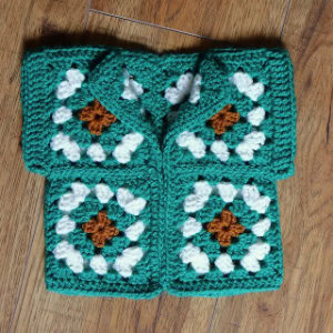 Un pull-over pour bébé fait à la main à partir de carrés de grand-mère crochetés en vert, blanc et marron, exposé sur un plancher en bois.