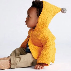 Un enfant en bas âge est assis sur le sol, vêtu d'une veste à capuche jaune texturée avec un pompon à l'extrémité, d'un pantalon beige et de chaussures couleur bronze, regardant sur le côté sur un fond uni.