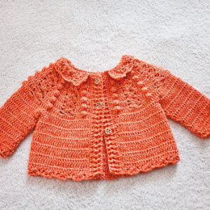 Un gilet pour bébé orange, crocheté à la main, avec des manches longues, un col festonné et trois boutons en bois, posé à plat sur une surface texturée de couleur claire.