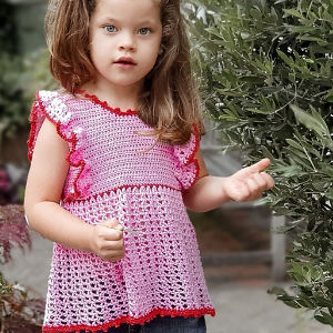 Une jeune fille aux longs cheveux bruns portant un haut crocheté rose avec des bordures rouges se tient à l'extérieur près d'un feuillage vert, regardant l'appareil photo avec une expression neutre.