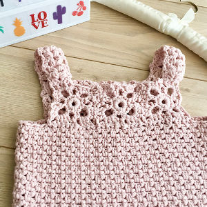 Une robe de bébé crochetée à la main, rose pâle, avec des motifs floraux sur le haut, présentée sur une surface en bois près d'une boîte contenant des autocollants colorés et un cintre blanc.