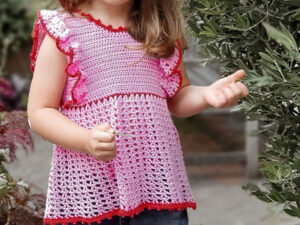 Un jeune enfant portant un modèle gratuit de top au crochet pour fille rose avec des bordures rouges se tient à l'extérieur près de plantes vertes, tenant un petit objet dans une main. L'enfant a de longs cheveux châtain clair et fait partiellement face à la caméra.