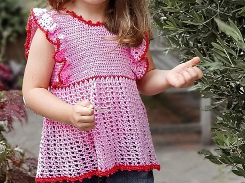 Un jeune enfant portant un modèle gratuit de top au crochet pour fille rose avec des bordures rouges se tient à l'extérieur près de plantes vertes, tenant un petit objet dans une main. L'enfant a de longs cheveux châtain clair et fait partiellement face à la caméra.