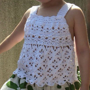 Un enfant portant un haut blanc au crochet, sans manches, avec un bord festonné et des motifs décoratifs, associé à une jupe verte et blanche. L'enfant est à l'extérieur.