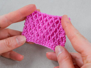 Deux mains tiennent un petit morceau rectangulaire de tissu rose crocheté, présentant un motif texturé de point nid d'abeille au crochet en forme de losange, sur une surface gris clair.