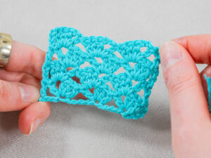 Mains tenant un petit morceau rectangulaire de tissu crocheté turquoise au motif ouvert et dentelle, mettant en valeur la technique délicate du point ajouré au crochet, sur un fond neutre.