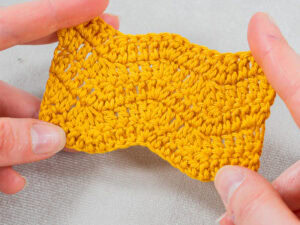 Gros plan de deux mains tenant un morceau de tissu jaune crocheté avec un motif au crochet ondulé et texturé en forme de chevron, sur un fond gris clair.