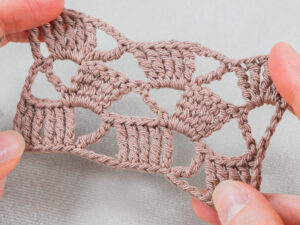 Gros plan de deux mains tenant un petit morceau de dentelle au crochet marron clair, présentant un motif complexe de crochet pour châle sur un fond neutre.