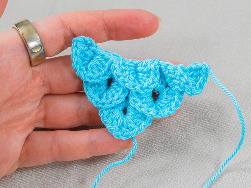 Une main avec un anneau d'argent tient un morceau de crochet bleu représentant un crocodile à pointe au crochet à motif festonné, sur un fond de tissu clair. Un bout de fil lâche s'échappe de la pièce crochetée.