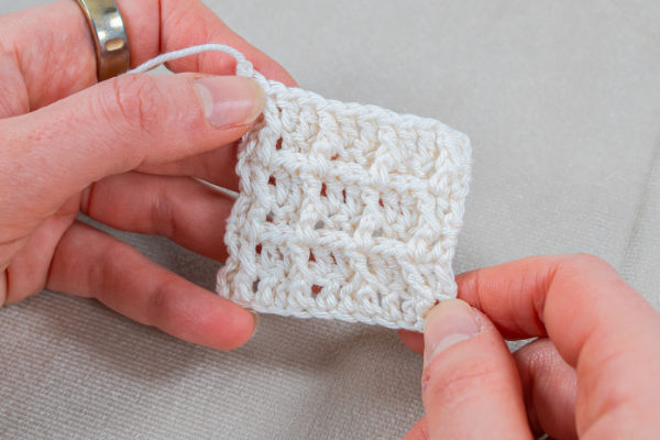 Une personne tient à deux mains un petit carré blanc au crochet représentant le motif du point gaufre au crochet. Une bague en argent est visible à un doigt sur un fond de tissu léger et texturé.