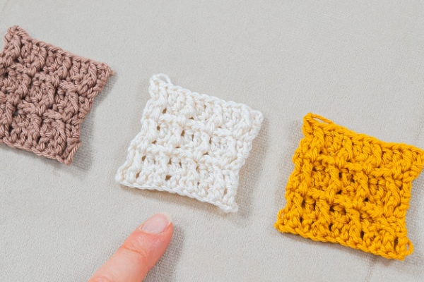 Trois petits carrés crochetés en marron, blanc (réalisé au point gaufré au crochet) et jaune sont placés côte à côte sur une surface claire. Un doigt pointe vers le carré blanc texturé au centre.