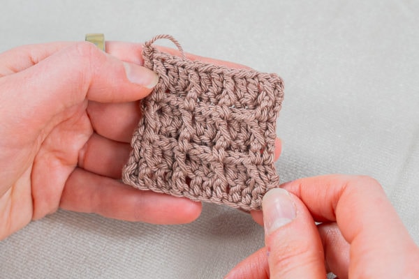 Deux mains tiennent un petit carré de tissu marron clair crocheté avec un motif géométrique texturé en point gaufré au crochet sur un fond neutre.