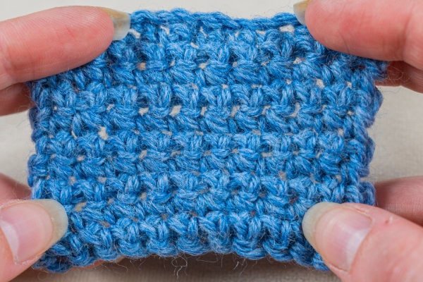 Gros plan sur des mains tenant un petit échantillon de tissu bleu au crochet, mettant en valeur la texture et les détails des mailles, réalisés selon la délicate technique du « point de rose au crochet ».