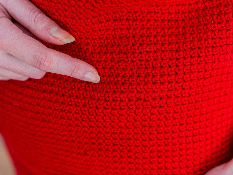 Une main à la peau pâle désigne un tissu au crochet d'un rouge vif, mettant en valeur la texture et le motif détaillés des mailles réalisées au point de rose au crochet.