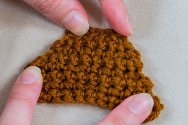 Deux mains tiennent une petite pièce au crochet, marron et texturée, de forme triangulaire — réalisée au point de rose au crochet — sur un fond de tissu beige.