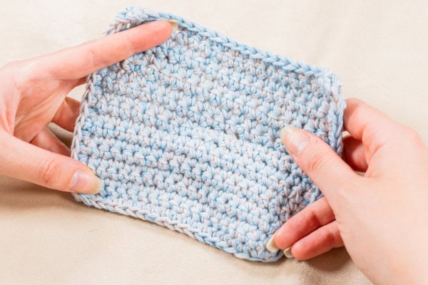 Deux mains tenant un petit échantillon rectangulaire au crochet, bleu et blanc, sur lequel se détache le délicat « point de rose au crochet » sur un fond clair.