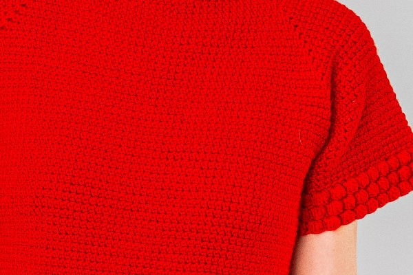 Gros plan sur une personne portant un pull en maille texturée rouge vif à manches courtes, orné de surpiqûres détaillées inspirées de la technique du « point de rose au crochet », sur fond gris.