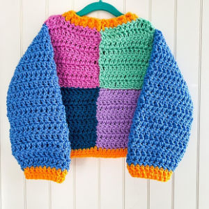 Un pull-over crocheté coloré à grosses mailles, avec des manches bleues, un corps divisé en carrés verts, roses, violets et bleus, et des bordures orange aux poignets, au col et à l'ourlet.