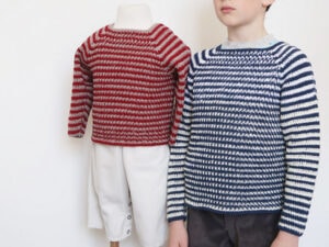 Un enfant portant un pull à rayures bleues et blanches, fabriqué à partir d'un tuto pull au crochet pour garcon, se tient à côté d'un mannequin vêtu d'un pull à rayures rouges et blanches et d'un pantalon blanc sur un fond blanc uni.