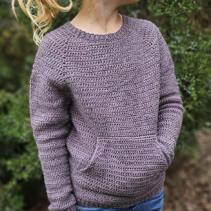 Une personne portant un pull en crochet violet clair fait à la main avec une poche avant se tient à l'extérieur devant des buissons verts.