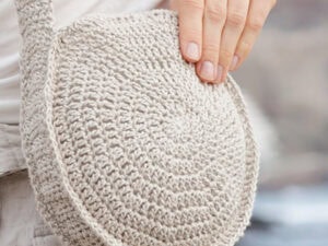 Une main tient un sac rond au crochet de couleur beige avec une lanière épaisse, porté en travers du corps. Le fond est flou et de couleur neutre, une inspiration parfaite pour votre prochain tuto sac rond au crochet en raphia.