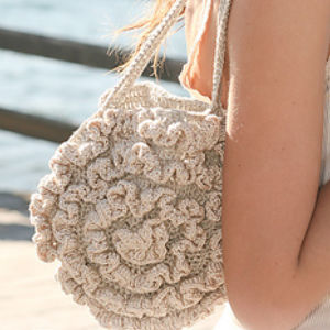 Une personne aux cheveux longs tient un sac rond, de couleur crème, crocheté avec des motifs floraux - un tuto sac rond au crochet en raphia - alors qu'elle se tient en plein air près d'un plan d'eau et d'une balustrade en bois.