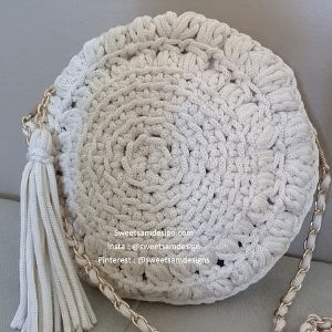 Un sac à main rond, blanc cassé, crocheté, doté d'un gros pompon et d'une lanière en chaîne dorée est présenté sur une surface de couleur claire. Le sac présente un fil épais et un motif circulaire étroitement tissé.