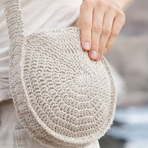 Une personne tient d'une main un sac à bandoulière rond, beige et crocheté. Le sac a un motif texturé en spirale et est assorti aux vêtements clairs de la personne.