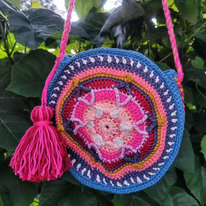 Un sac rond coloré, crocheté, avec un pompon et une lanière roses, est suspendu devant des plantes vertes et feuillues. Le sac présente des motifs concentriques en fil bleu, rose, crème, blanc et jaune.