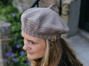 Femme aux longs cheveux blonds portant un béret texturé marron clair femme au crochet, en plein air près d'un mur de pierre et de fleurs violettes.