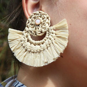 Gros plan sur l'oreille d'une personne portant une grande boucle d'oreille circulaire en paille tressée avec une frange en forme d'éventail et une petite perle au centre.