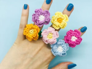Une main aux ongles peints en bleu porte six anneaux de fleurs au crochet colorés -bijoux au crochet- en jaune, violet, rose, bleu et orange sur un fond bleu clair.