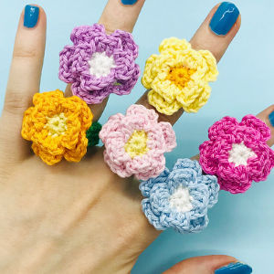 Une main aux ongles peints en bleu porte cinq anneaux de fleurs colorées au crochet, chacun dans une nuance pastel différente, sur un fond bleu clair.
