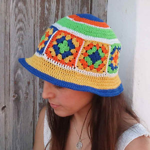 Une femme portant un chapeau coloré au crochet avec des motifs carrés de grand-mère en vert, bleu, orange et jaune, se tient devant une porte en bois rustique.
