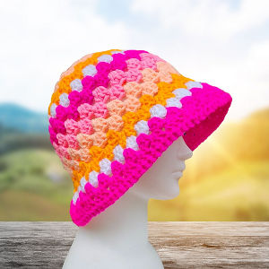 La tête d'un mannequin blanc est coiffée d'un chapeau de seau en crochet aux rayures roses, orange, jaunes et blanches, sur un fond flou de soleil et de collines verdoyantes.