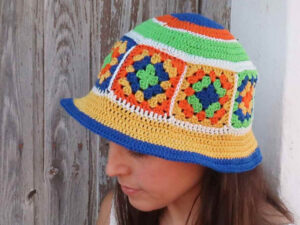 Une femme portant un chapeau en crochet coloré avec des motifs géométriques en bleu, vert, orange, jaune et blanc se tient près d'un mur en bois.
